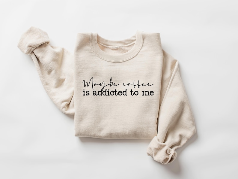 May include: Cream-colored sweatshirt with the text "Maybe coffee is addicted to me" in black script. The sleeves are rolled up, and the garment is laid flat. A casual, comfortable apparel item.