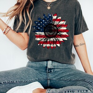 May include: Dark gray t-shirt featuring a sunflower design with the American flag's colors. The sunflower's petals are red and white stripes, and the center is a blue field with white stars. The shirt is paired with blue jeans and white sneakers.