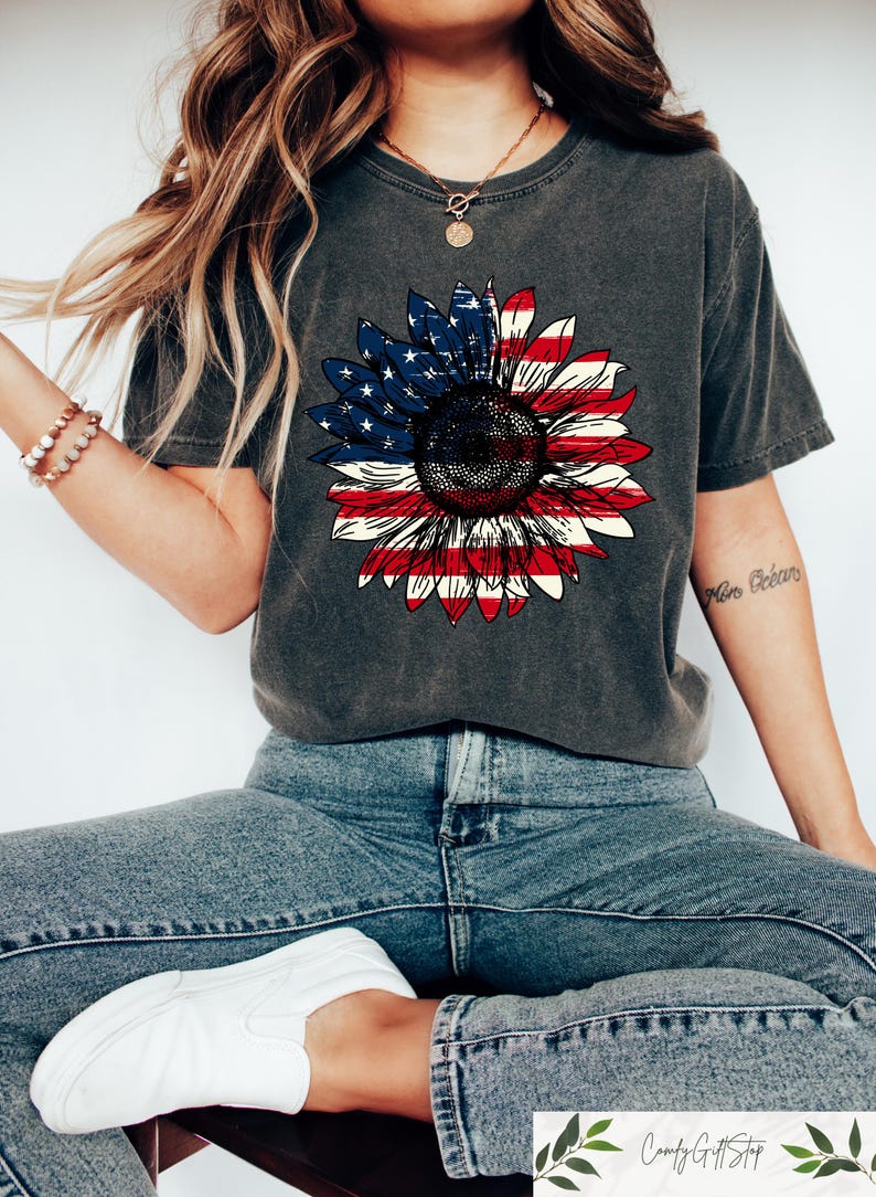 May include: Dark gray t-shirt featuring a sunflower design with the American flag's colors. The sunflower's petals are red and white stripes, and the center is a blue field with white stars. The shirt is paired with blue jeans and white sneakers.