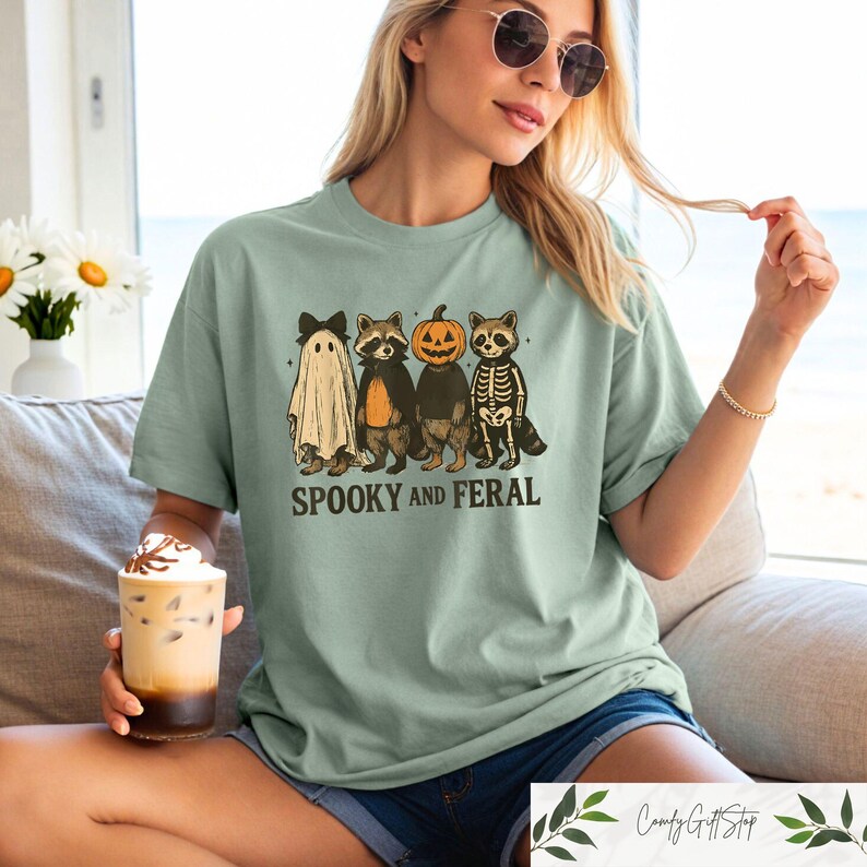 May include: Sage green t-shirt with a Halloween graphic. The design features a ghost, two raccoons, and a pumpkin, with the text "Spooky and Feral" underneath. The t-shirt has a loose, comfortable fit.