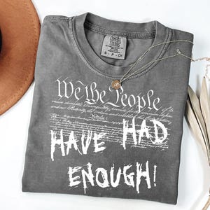Puede incluir: Una camiseta gris Comfort Colors con el texto "We the People HAVE HAD ENOUGH!" en blanco. Un collar dorado y un sombrero marr&oacute;n tambi&eacute;n est&aacute;n en la imagen.