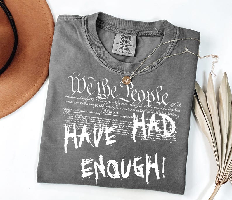 Puede incluir: Una camiseta gris Comfort Colors con el texto "We the People HAVE HAD ENOUGH!" en blanco. Un collar dorado y un sombrero marr&oacute;n tambi&eacute;n est&aacute;n en la imagen.