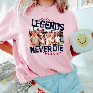 May include: A light pink t-shirt featuring a graphic of a baseball and a group of boys with the text "LEGENDS NEVER DIE". The shirt is a crew neck style.