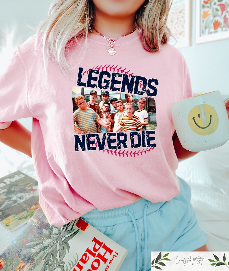 May include: A light pink t-shirt featuring a graphic of a baseball and a group of boys with the text "LEGENDS NEVER DIE". The shirt is a crew neck style.
