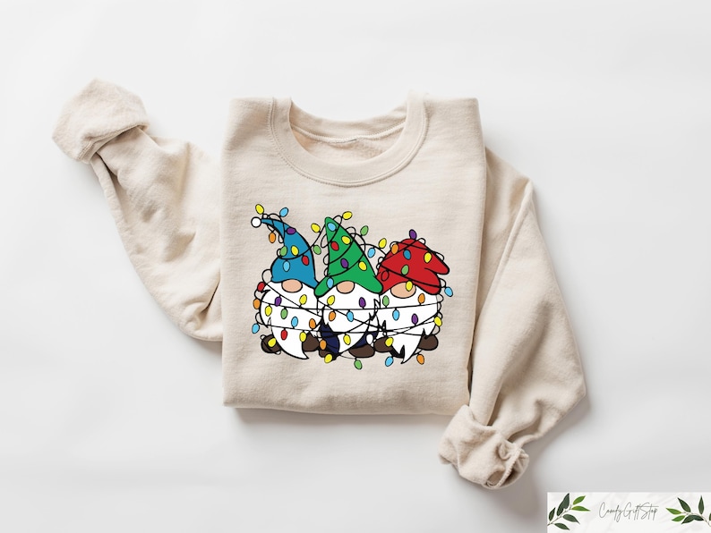 May include: Beige sweatshirt featuring a graphic of three gnomes wrapped in Christmas lights. The gnomes wear blue, green, and red hats. The sweatshirt has long sleeves and a crew neck.