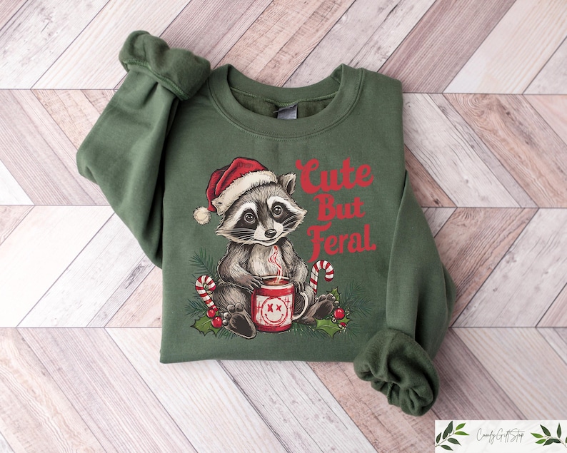 Pu&ograve; includere: Felpa verde natalizia con un adorabile procione che indossa un cappello da Babbo Natale e tiene una tazza di cioccolata calda. Il testo "Cute But Feral" &egrave; stampato sulla felpa.