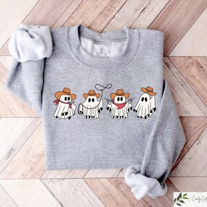May include: A grey sweatshirt featuring a graphic of five ghosts dressed as cowboys. The ghosts are wearing cowboy hats, bandanas, and holsters. The text "Boo Crew" is printed on the sweatshirt.