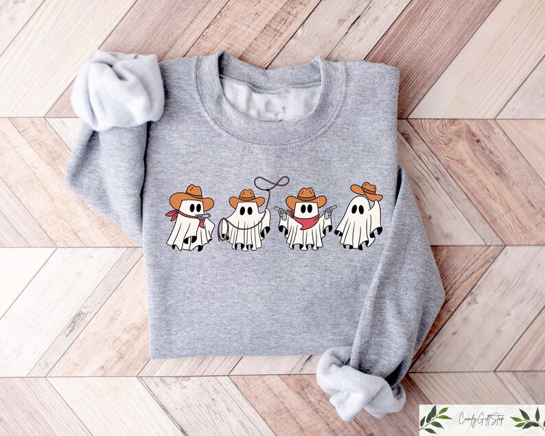 May include: A grey sweatshirt featuring a graphic of five ghosts dressed as cowboys. The ghosts are wearing cowboy hats, bandanas, and holsters. The text "Boo Crew" is printed on the sweatshirt.