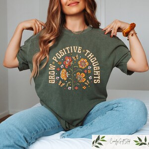 Pu&ograve; includere: Una maglietta verde con un design floreale vintage e il testo "Grow Positive Thoughts" in un cerchio attorno ai fiori.