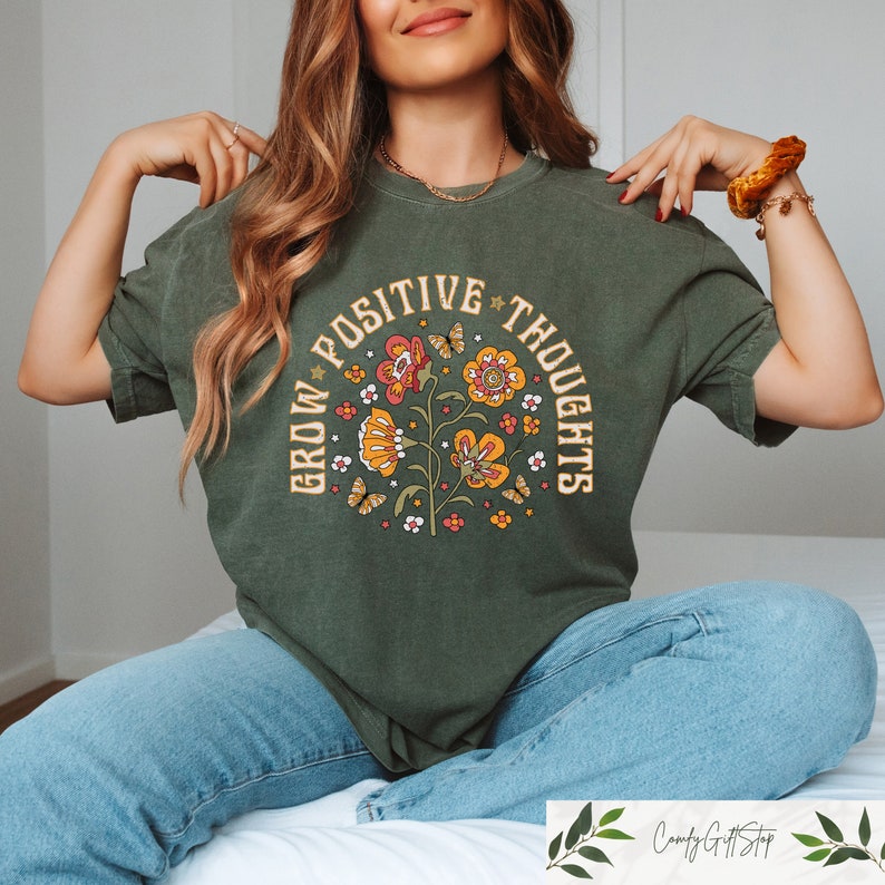 Pu&ograve; includere: Una maglietta verde con un design floreale vintage e il testo "Grow Positive Thoughts" in un cerchio attorno ai fiori.