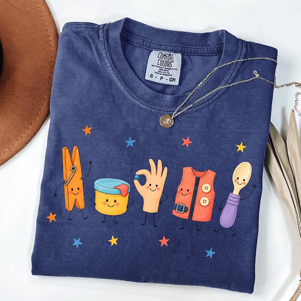 May include: A navy blue short-sleeve t-shirt with a Comfort Colors label. The graphic features five cartoon characters: an orange clothespin, a yellow container, a hand making an okay sign, an orange vest, and a spoon, surrounded by small stars.