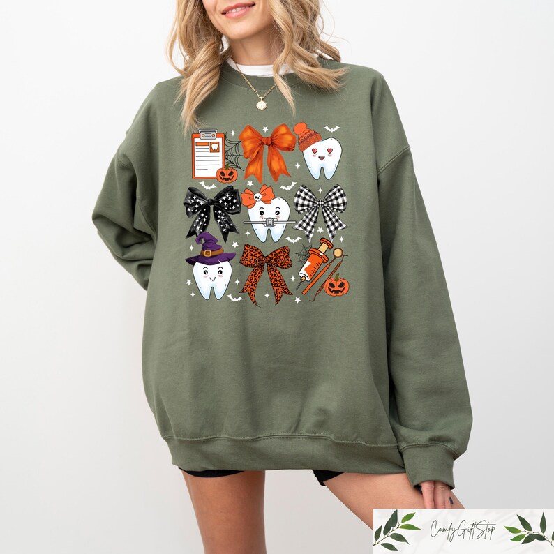 May include: A green sweatshirt featuring a Halloween-themed dental design. The design includes teeth with bows, a witch's hat, a syringe, and a clipboard with a tooth on it.