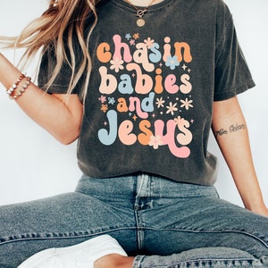 Peut inclure: Un t-shirt gris fonc&eacute; avec un graphisme r&eacute;tro qui dit "chasin babies and Jesus" en lettres color&eacute;es avec des fleurs et des &eacute;toiles autour du texte.