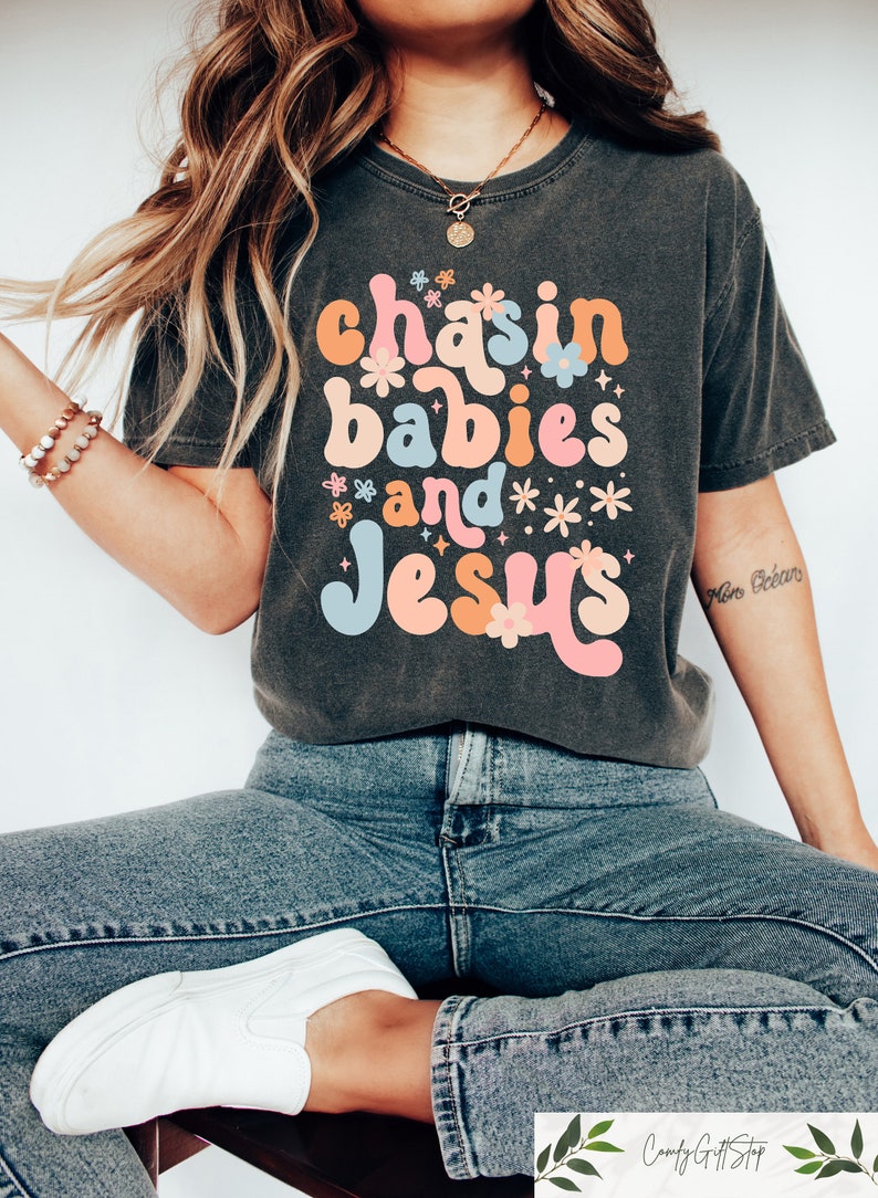 Peut inclure: Un t-shirt gris fonc&eacute; avec un graphisme r&eacute;tro qui dit "chasin babies and Jesus" en lettres color&eacute;es avec des fleurs et des &eacute;toiles autour du texte.