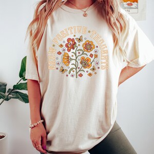 Pu&ograve; includere: Una maglietta color crema con una grafica di un fiore con farfalle e stelle. Il testo "Grow Positive Thoughts" &egrave; scritto in un cerchio attorno al fiore.