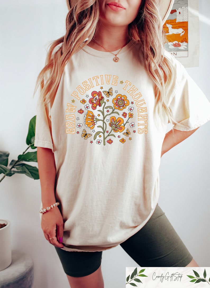 Pu&ograve; includere: Una maglietta color crema con una grafica di un fiore con farfalle e stelle. Il testo "Grow Positive Thoughts" &egrave; scritto in un cerchio attorno al fiore.