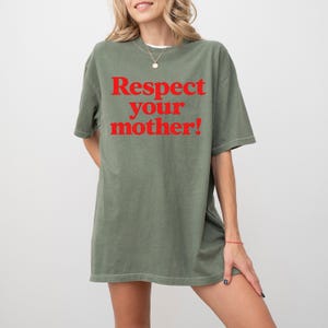 May include: An olive green t-shirt with the red text "Respect your mother!". The shirt is a loose fit and appears to be made of a soft material. The model is wearing a necklace and a bracelet.