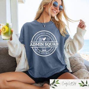 Pu&ograve; includere: Una maglietta blu con una grafica circolare bianca che dice "Organizzare il caos da sempre ADMIN SQUAD WE SLAY ALL DAY". La grafica include icone di una lista di controllo, un cuore, un archivio e un laptop.