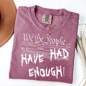 Puede incluir: Una camiseta de color malva con el texto blanco "We the People HAVE HAD ENOUGH!". La camiseta tiene cuello redondo y una peque&ntilde;a etiqueta en el cuello. Un collar dorado con un colgante est&aacute; sobre la camiseta.