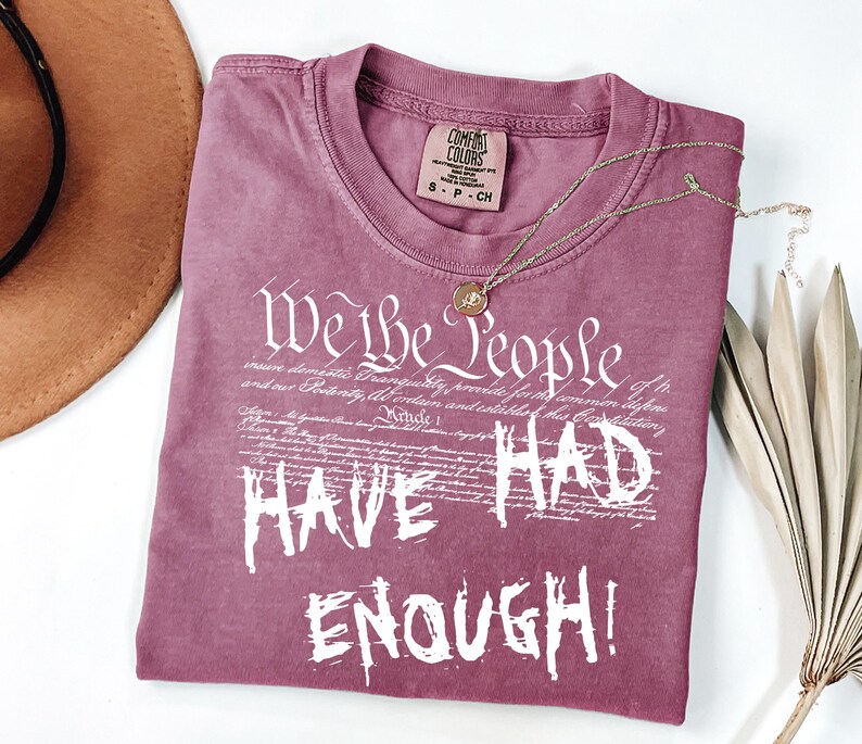 Puede incluir: Una camiseta de color malva con el texto blanco "We the People HAVE HAD ENOUGH!". La camiseta tiene cuello redondo y una peque&ntilde;a etiqueta en el cuello. Un collar dorado con un colgante est&aacute; sobre la camiseta.