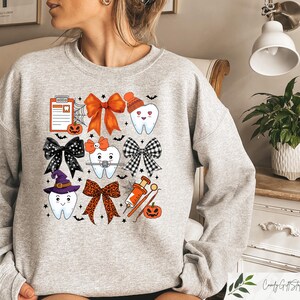 May include: A grey sweatshirt with a Halloween-themed design featuring cartoon teeth with bows, a witch's hat, and a dental tool. The design also includes a pumpkin, a spiderweb, and a clipboard with a tooth icon.
