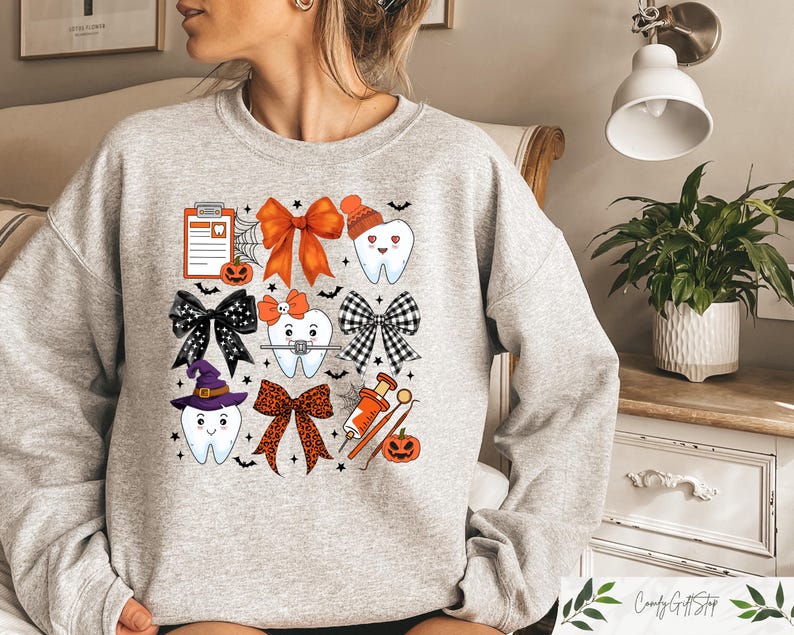 May include: A grey sweatshirt with a Halloween-themed design featuring cartoon teeth with bows, a witch's hat, and a dental tool. The design also includes a pumpkin, a spiderweb, and a clipboard with a tooth icon.