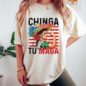 May include: Cream-colored t-shirt featuring a graphic of a woman in a sombrero flexing her arm, with the text "CHINGA TU MAGA" in black and red. The design incorporates the American flag and a rose.