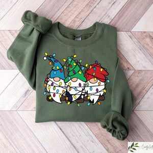 May include: An olive green sweatshirt featuring a graphic of three gnomes wearing colorful hats and wrapped in Christmas lights. The gnomes have white beards and are smiling. The sweatshirt has long sleeves and a crew neck.