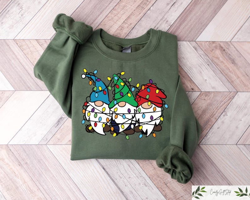 May include: An olive green sweatshirt featuring a graphic of three gnomes wearing colorful hats and wrapped in Christmas lights. The gnomes have white beards and are smiling. The sweatshirt has long sleeves and a crew neck.