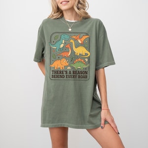 May include: Olive green t-shirt featuring a colorful dinosaur graphic. The graphic includes various dinosaurs in shades of blue, orange, and yellow, with the text "THERE'S A REASON BEHIND EVERY ROAR" in a rectangular frame. The shirt has short sleeves.