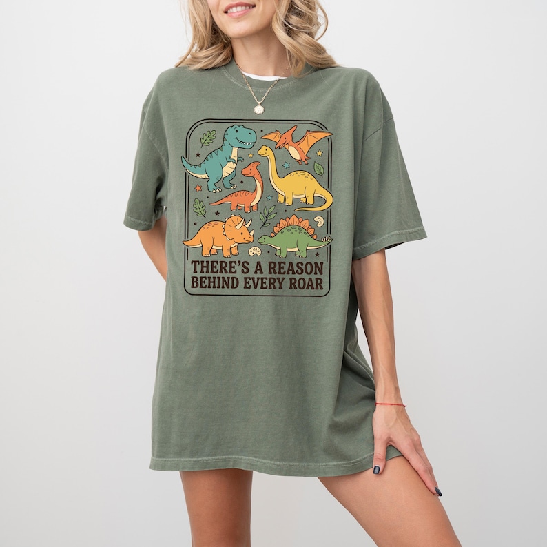May include: Olive green t-shirt featuring a colorful dinosaur graphic. The graphic includes various dinosaurs in shades of blue, orange, and yellow, with the text "THERE'S A REASON BEHIND EVERY ROAR" in a rectangular frame. The shirt has short sleeves.