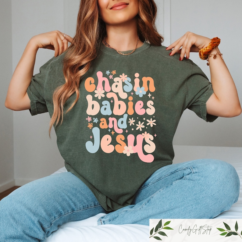 Peut inclure: Un t-shirt vert avec le texte "chasin babies and Jesus" dans une police r&eacute;tro avec des fleurs color&eacute;es entourant le texte.
