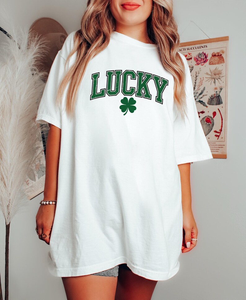 May include: White t-shirt featuring the word "LUCKY" in green, block lettering, and a green four-leaf clover. The shirt has a relaxed, oversized fit, suitable for St. Patrick's Day or casual wear.