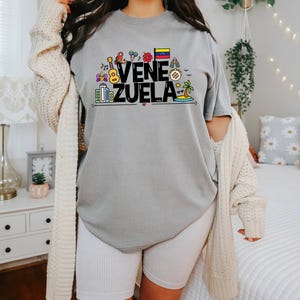 May include: A gray t-shirt with a black graphic design that says "VENEZUELA" with colorful icons representing Venezuelan culture.
