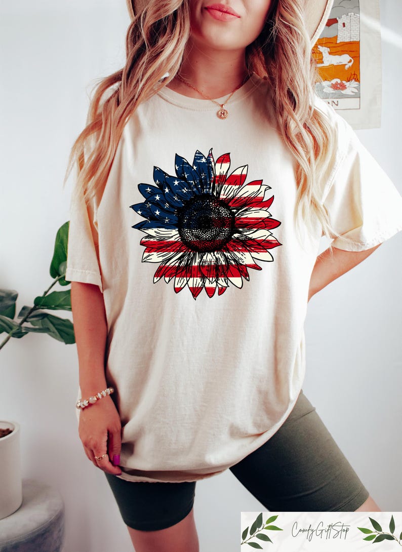 May include: Cream-colored t-shirt featuring a large sunflower design. The sunflower's petals are colored with the American flag, red and white stripes on the lower half and blue with white stars on the upper half. The shirt is a casual style.