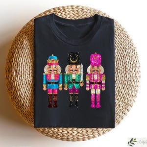 May include: Black t-shirt with three glittery nutcracker figures. The nutcrackers are wearing blue, green, and pink outfits with gold accents.