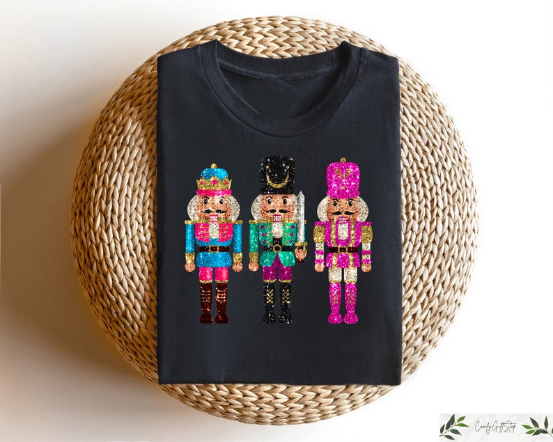 May include: Black t-shirt with three glittery nutcracker figures. The nutcrackers are wearing blue, green, and pink outfits with gold accents.