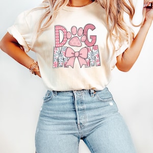 May include: Cream-colored t-shirt with the text "DOG MOM" in pink and floral designs. The "D" and "G" are pink with white polka dots and stripes, respectively. A paw print and bow are also featured.