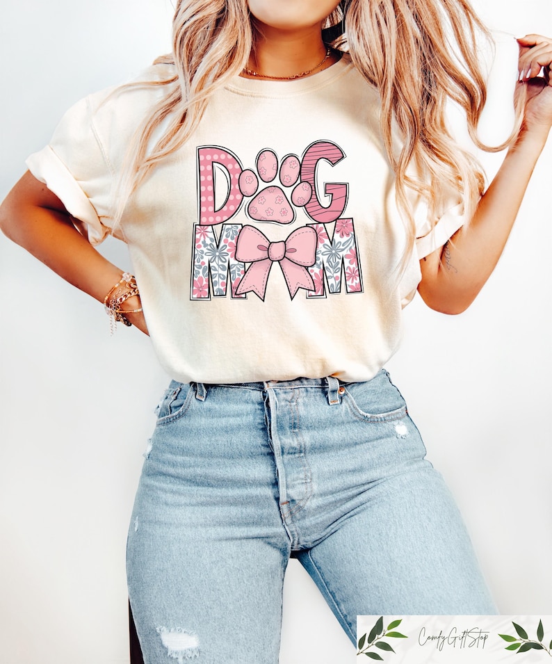 May include: Cream-colored t-shirt with the text "DOG MOM" in pink and floral designs. The "D" and "G" are pink with white polka dots and stripes, respectively. A paw print and bow are also featured.