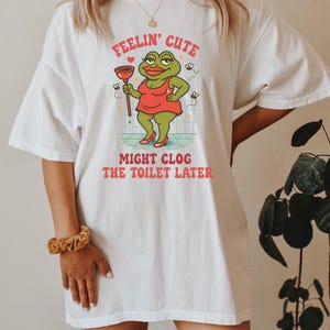 以下が含まれることがあります： 白いTシャツに漫画の蛙のデザイン。蛙は赤いドレスを着て、ラバーカップを持っています。Tシャツのテキストは「Feelin' Cute Might Clog The Toilet Later」と書かれています。