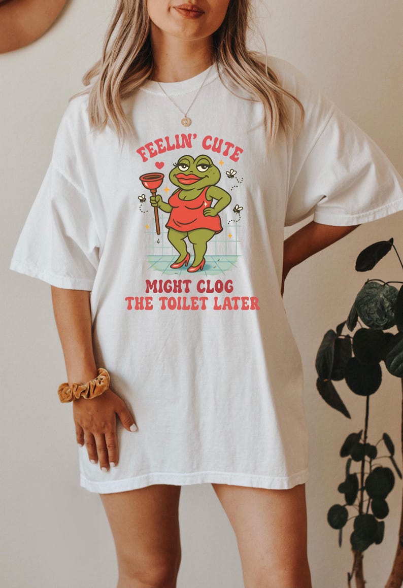 以下が含まれることがあります： 白いTシャツに漫画の蛙のデザイン。蛙は赤いドレスを着て、ラバーカップを持っています。Tシャツのテキストは「Feelin' Cute Might Clog The Toilet Later」と書かれています。