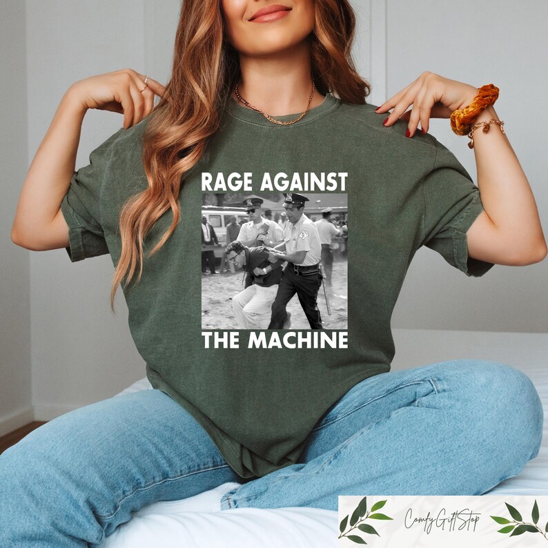 May include: Olive green t-shirt featuring a black and white image of a protest scene with the text "RAGE AGAINST THE MACHINE". The shirt is made of a soft material and has short sleeves.