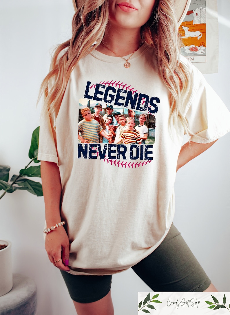 May include: Cream-colored t-shirt featuring a group photo and the text "LEGENDS NEVER DIE" in navy blue. The design includes a baseball and stitching. The shirt is a relaxed fit.