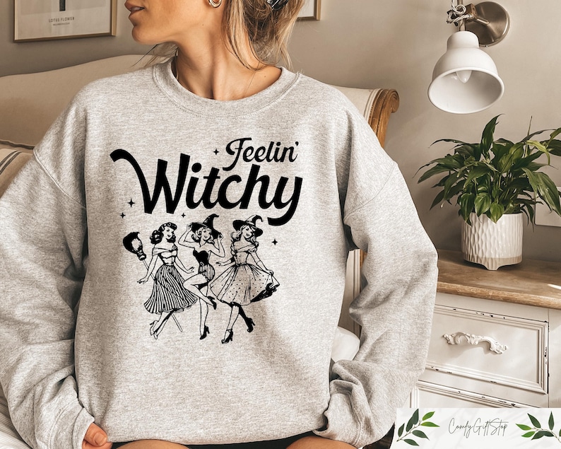 Puede incluir: Sudadera gris claro con la frase "Feelin' Witchy" en negro sobre un gr&aacute;fico de tres brujas bailando. Las brujas est&aacute;n en blanco y negro, con vestidos y sombreros de bruja. La sudadera es de cuello redondo.