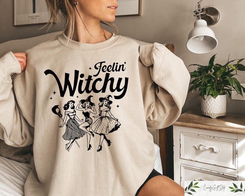 Puede incluir: Sudadera beige con la frase "Feelin' Witchy" en letras negras. Debajo, un gr&aacute;fico de tres brujas bailando con vestidos y sombreros. La sudadera es de color neutro.