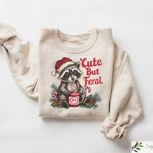 Pu&ograve; includere: Una felpa beige con una grafica natalizia di un procione che indossa un cappello da Babbo Natale e tiene una tazza. Il testo "Cute But Feral" &egrave; stampato in rosso sulla felpa.