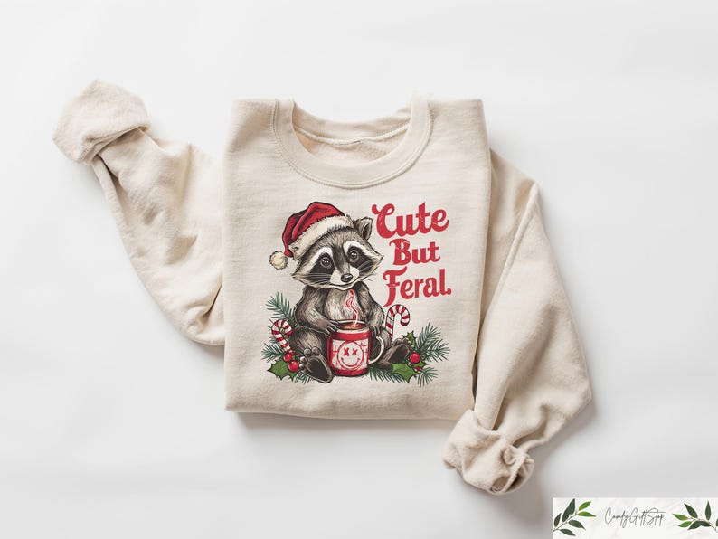 Pu&ograve; includere: Una felpa beige con una grafica natalizia di un procione che indossa un cappello da Babbo Natale e tiene una tazza. Il testo "Cute But Feral" &egrave; stampato in rosso sulla felpa.