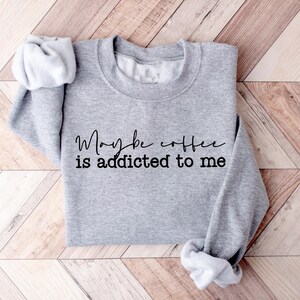 May include: A heather gray sweatshirt with the phrase "Maybe coffee is addicted to me" in black script. The sweatshirt has a crew neck and long sleeves, with the sleeves folded up. The garment is displayed on a wooden surface.