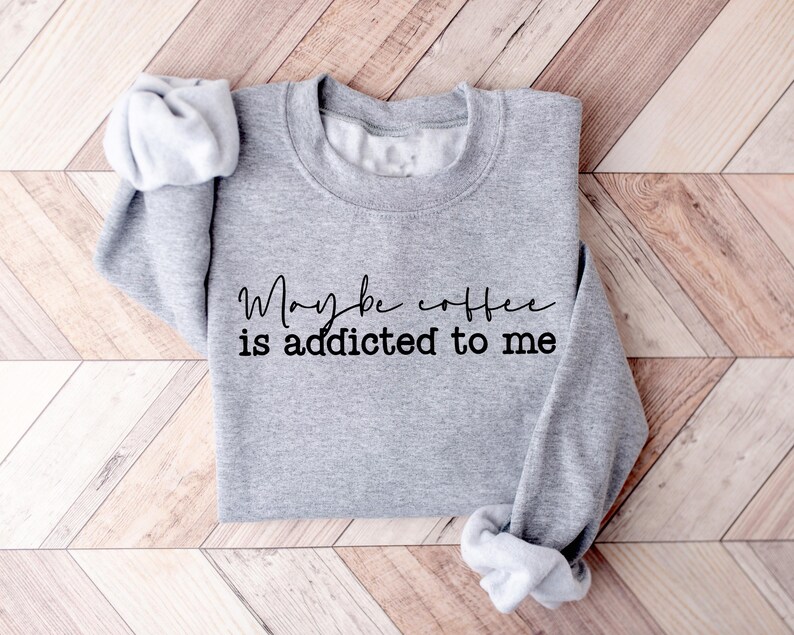 May include: A heather gray sweatshirt with the phrase "Maybe coffee is addicted to me" in black script. The sweatshirt has a crew neck and long sleeves, with the sleeves folded up. The garment is displayed on a wooden surface.