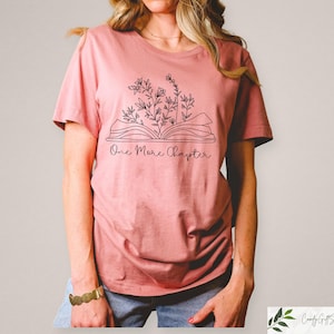 May include: A pink t-shirt with a black line drawing of an open book with flowers growing out of it. The text "One More Chapter" is printed below the book.